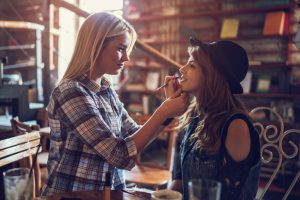 Young woman preparing her female friend for a date in a cafe. She is applying lip balm on her mouth.